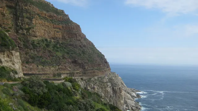 A narrow road follows a layered coastal cliff above blue sea.