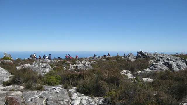 Visitors gather along a rocky plateau beneath a blue ocean horizon.