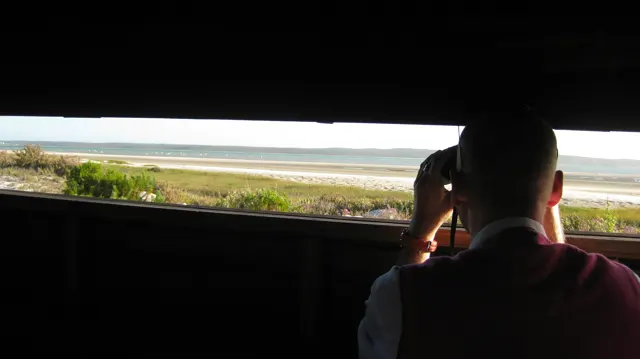 A silhouetted visitor looks through binoculars from a dark bird hide.