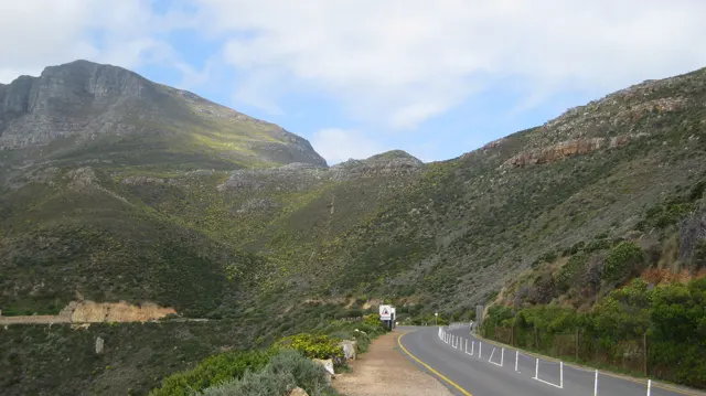 A road curves between green mountain slopes beside white posts.