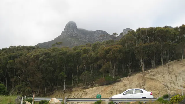 A white car passes below trees and a steep rocky peak.