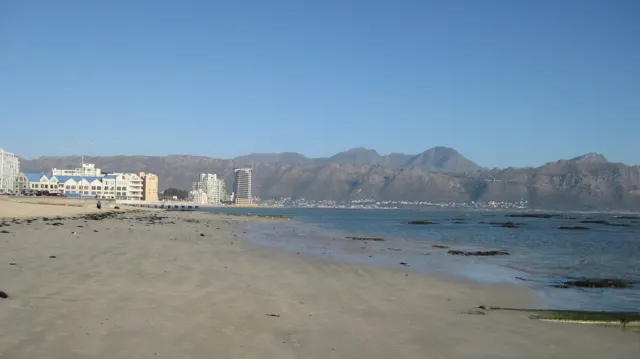A pale beach faces a long rocky mountain range across water.