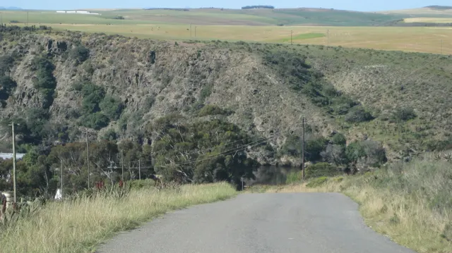 A narrow road descends beside grass toward a steep valley wall.