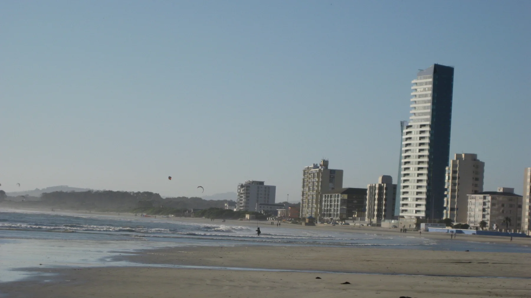 A narrow dark-topped tower rises above waterfront buildings and shallow surf.