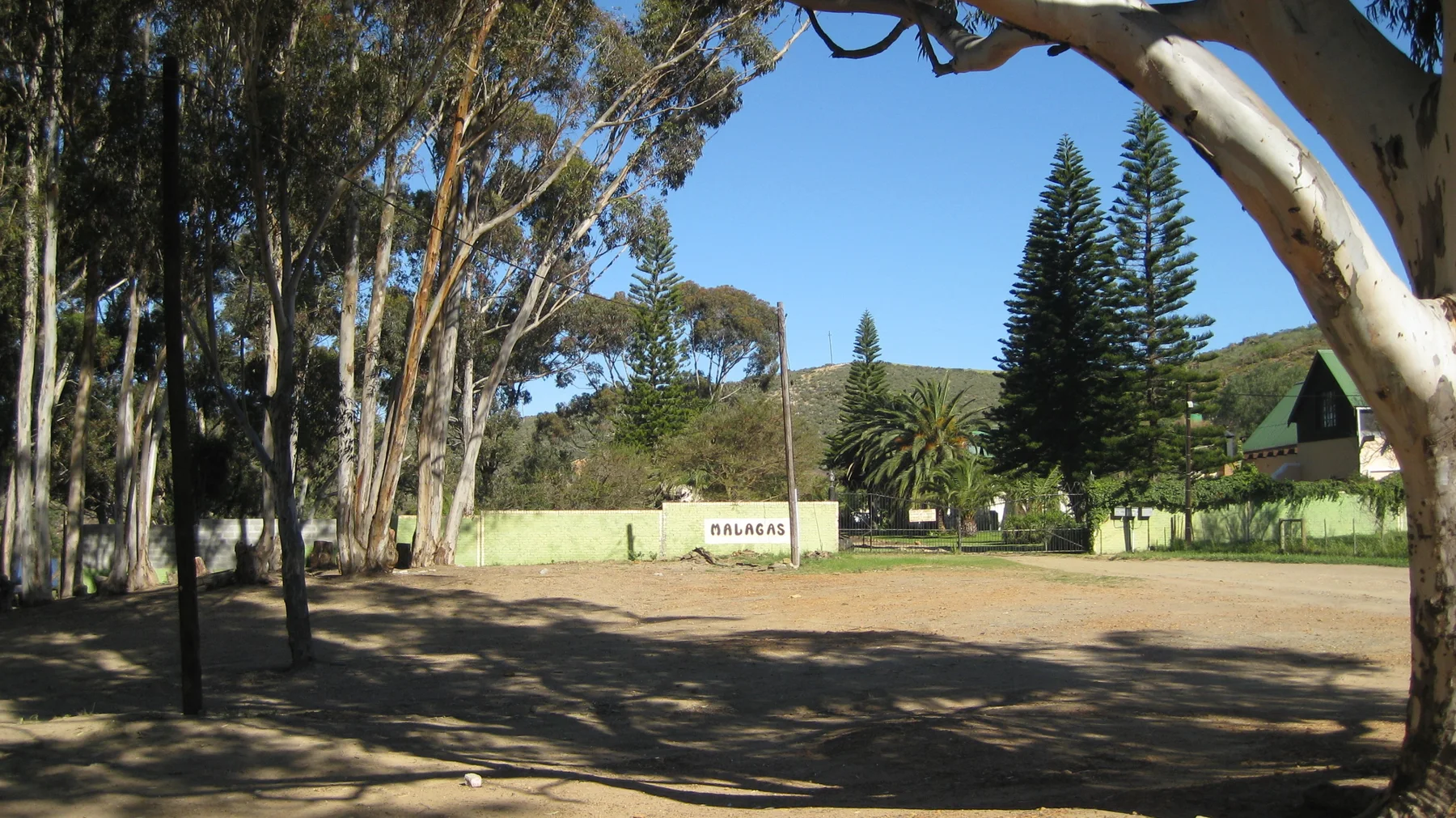 Tall trees shade an entrance beside pale walls and green grounds.