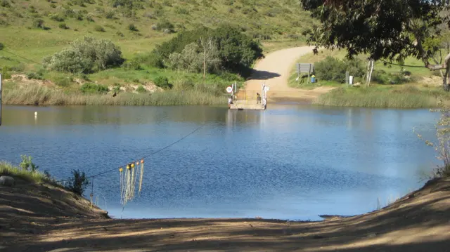 A dirt road reaches a calm blue river with a landing opposite.