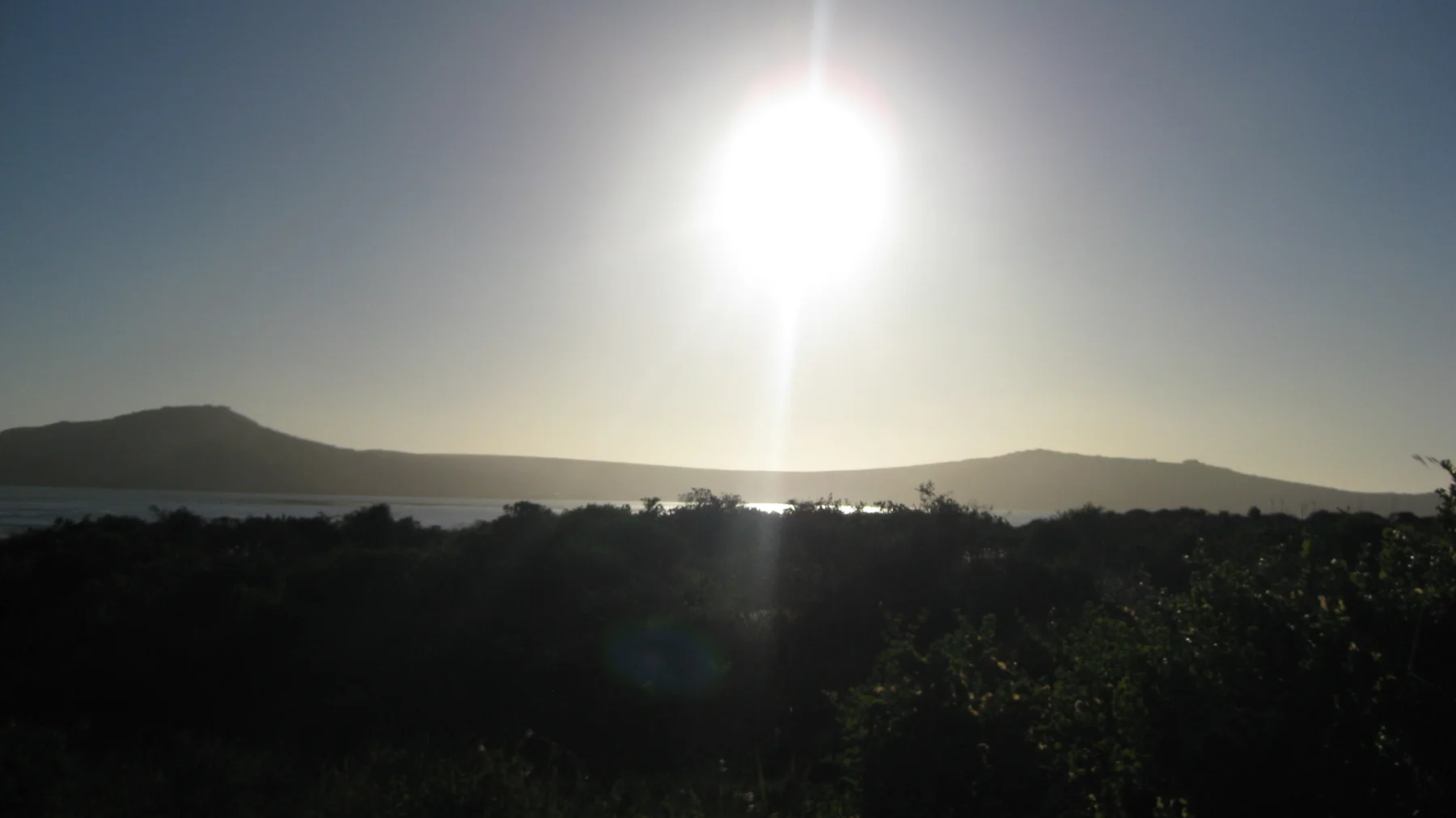 A bright sun hangs above silhouetted land and reflective water.