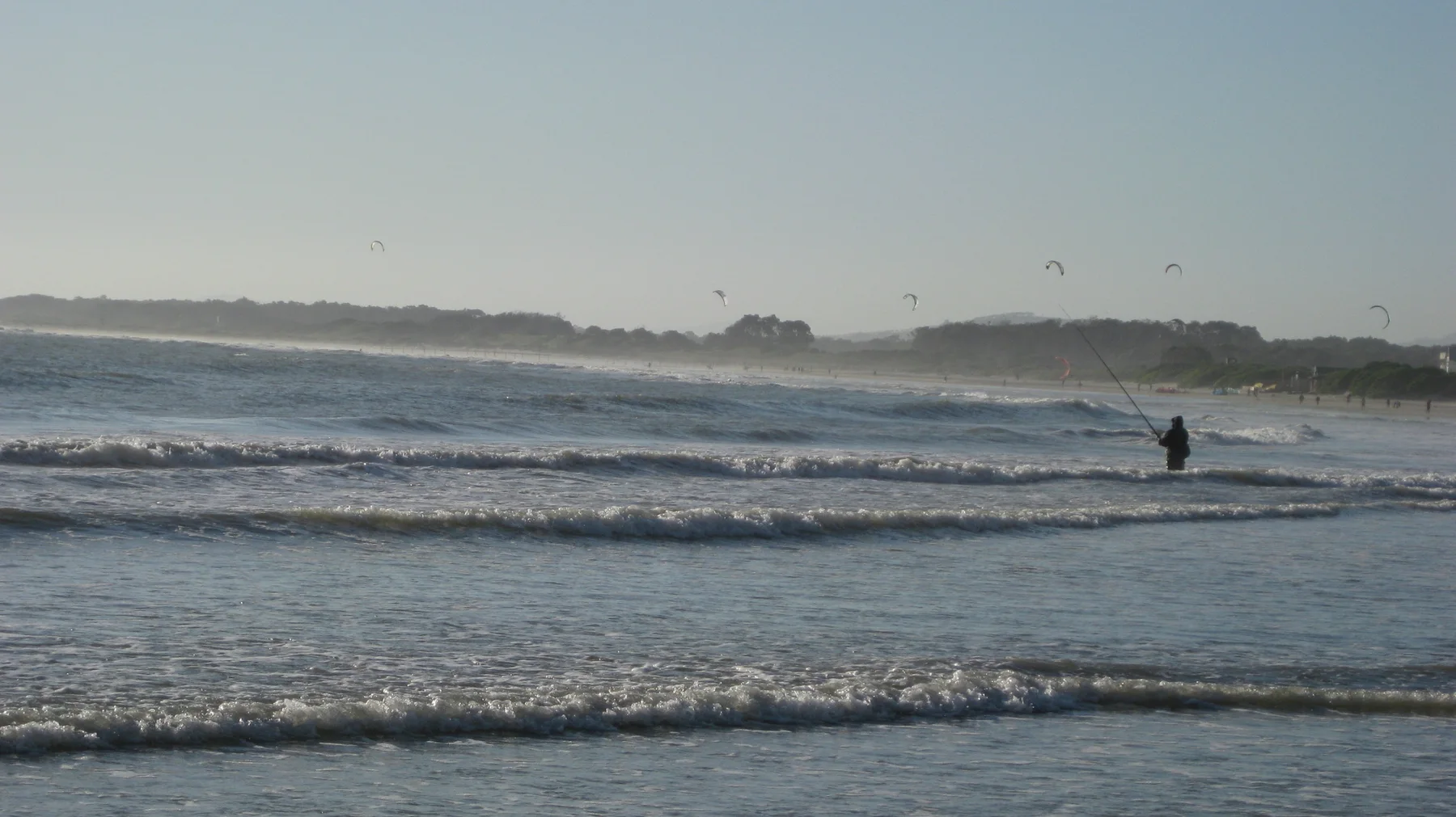 A lone figure stands in shallow surf beneath a hazy shoreline.