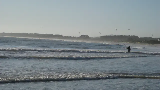 A lone figure stands in shallow surf beneath a hazy shoreline.