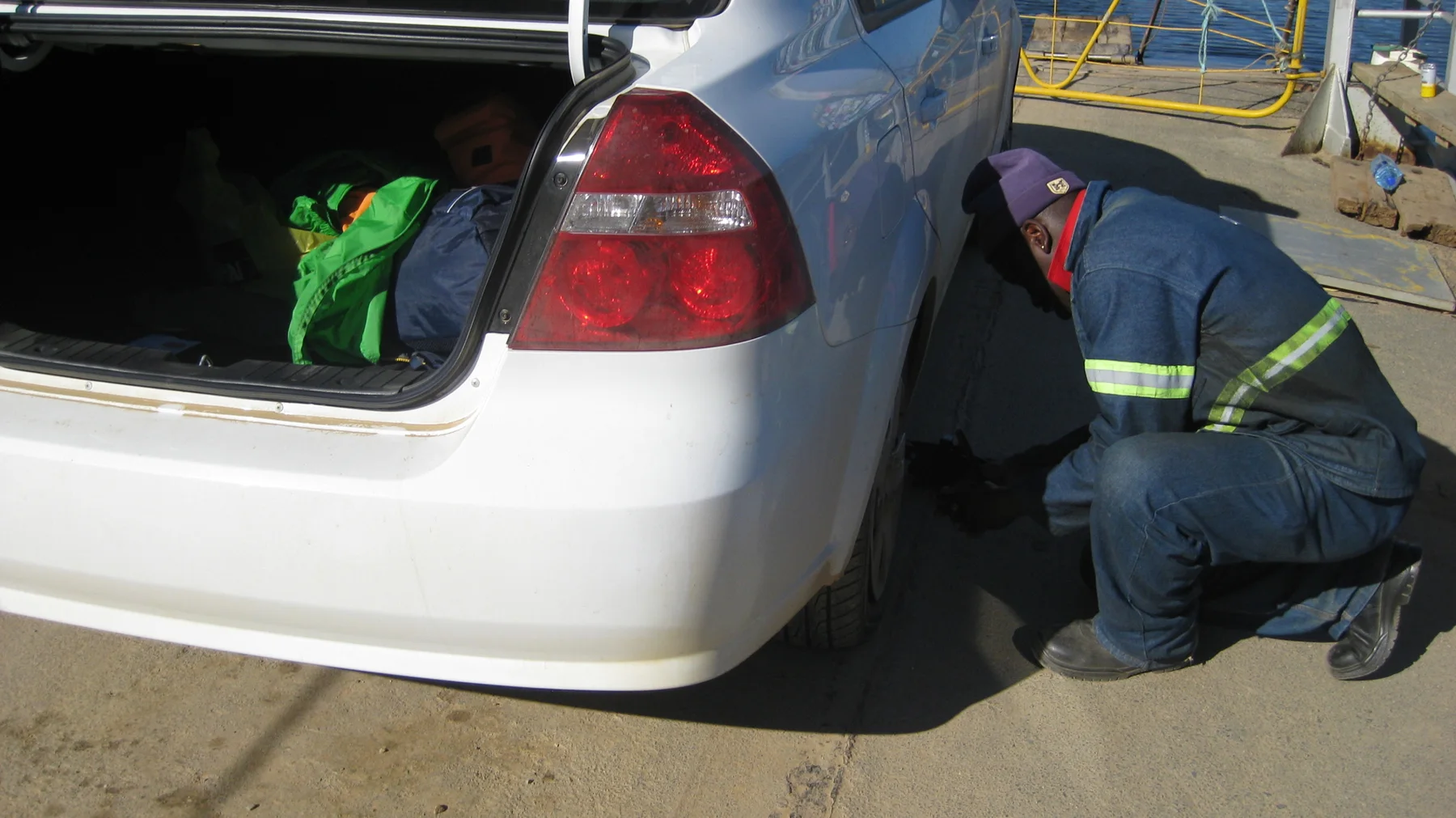 A person crouches beside a car's rear wheel beneath an open trunk.