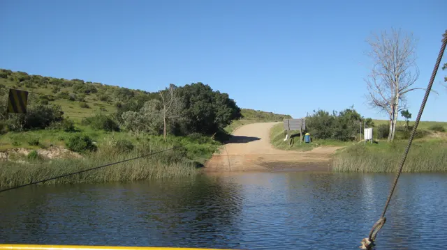 A dirt road descends to the opposite riverbank beside reeds.