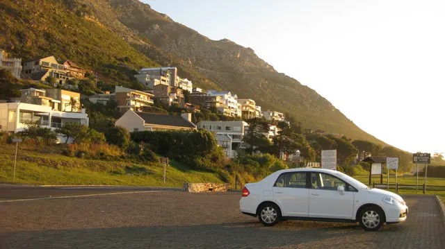 A white car stands beside a road below sunlit hillside houses.
