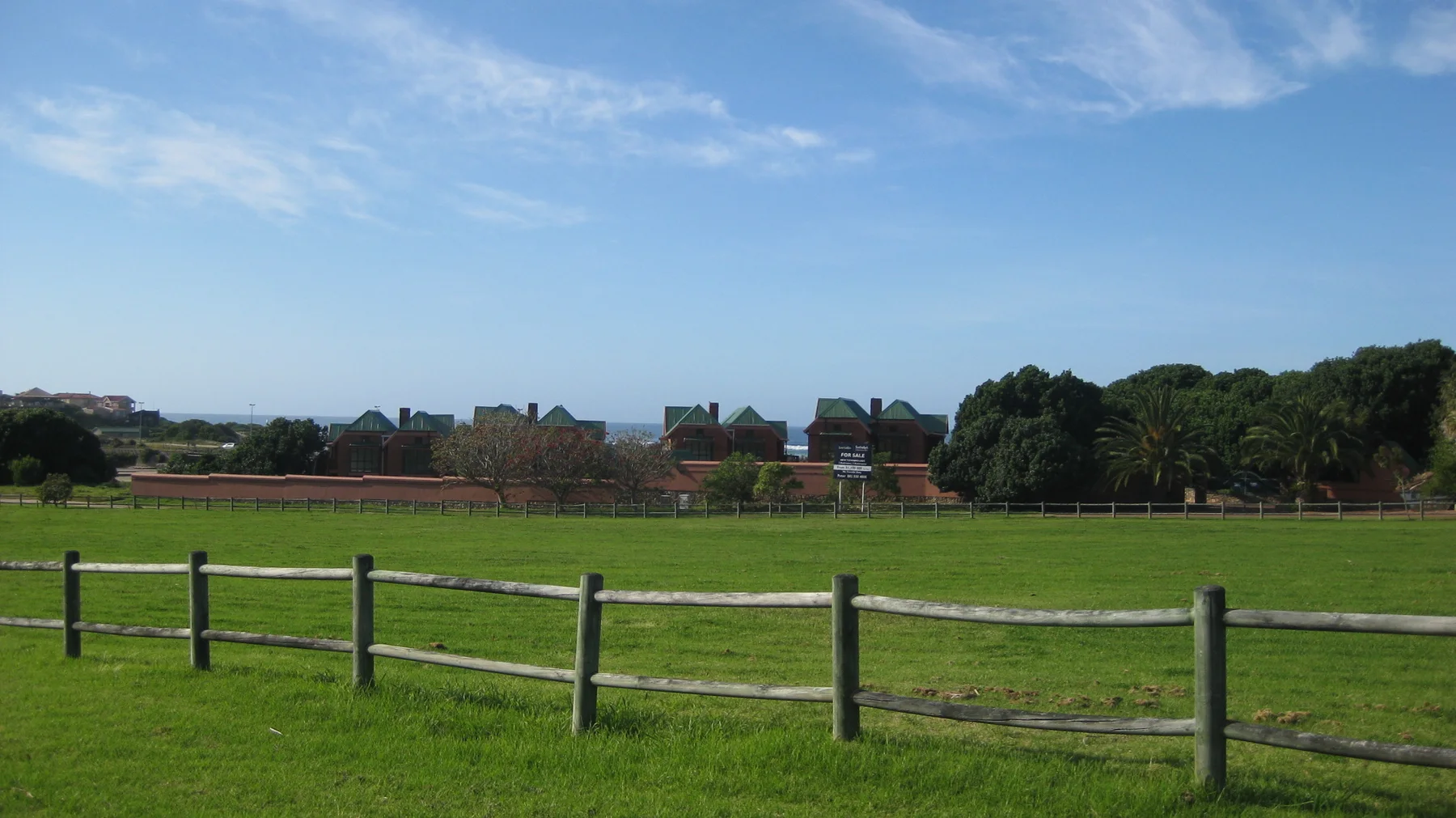 A wooden fence borders a wide green field and distant buildings.