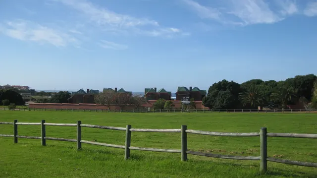 A wooden fence borders a wide green field and distant buildings.