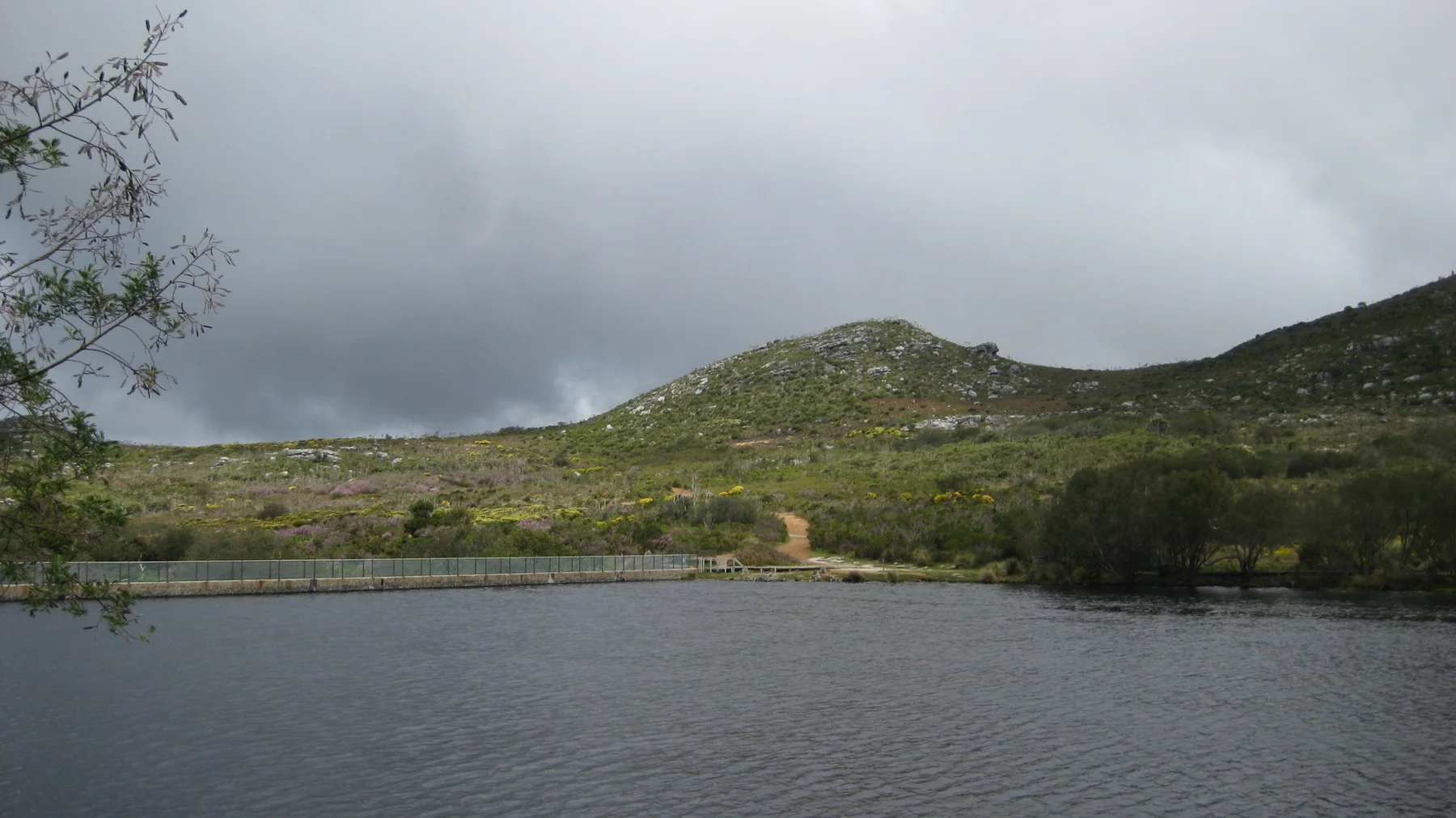 Dark clouds gather above a reservoir and low green hills.