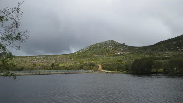 Dark clouds gather above a reservoir and low green hills.