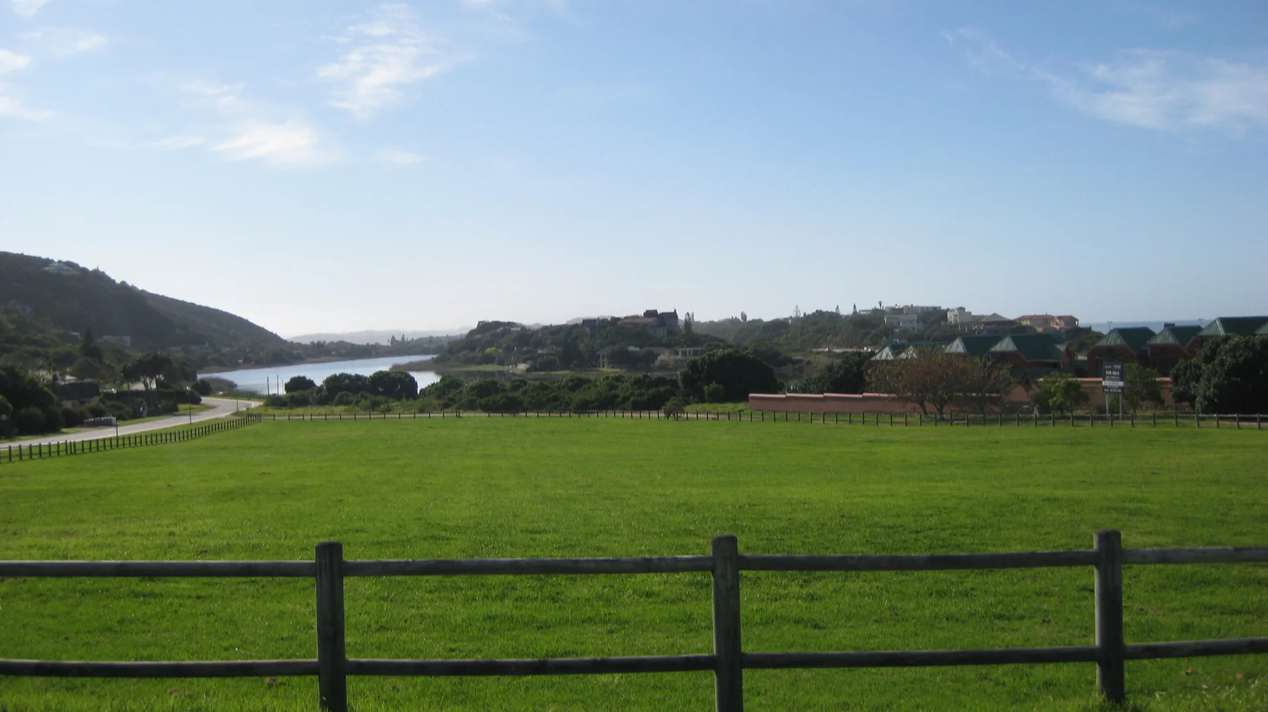 A broad green field leads toward coastal buildings and distant water.