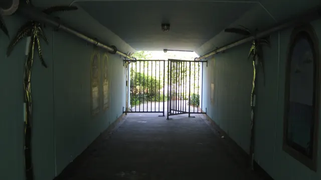 A dim covered passage leads toward a bright metal gate.