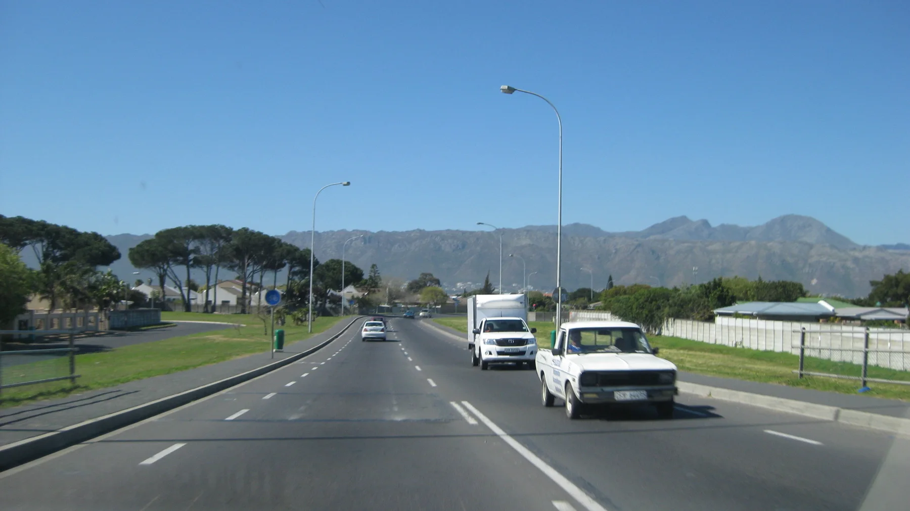 Cars pass along a broad road below a long mountain ridge.