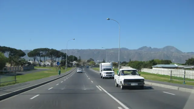 Cars pass along a broad road below a long mountain ridge.