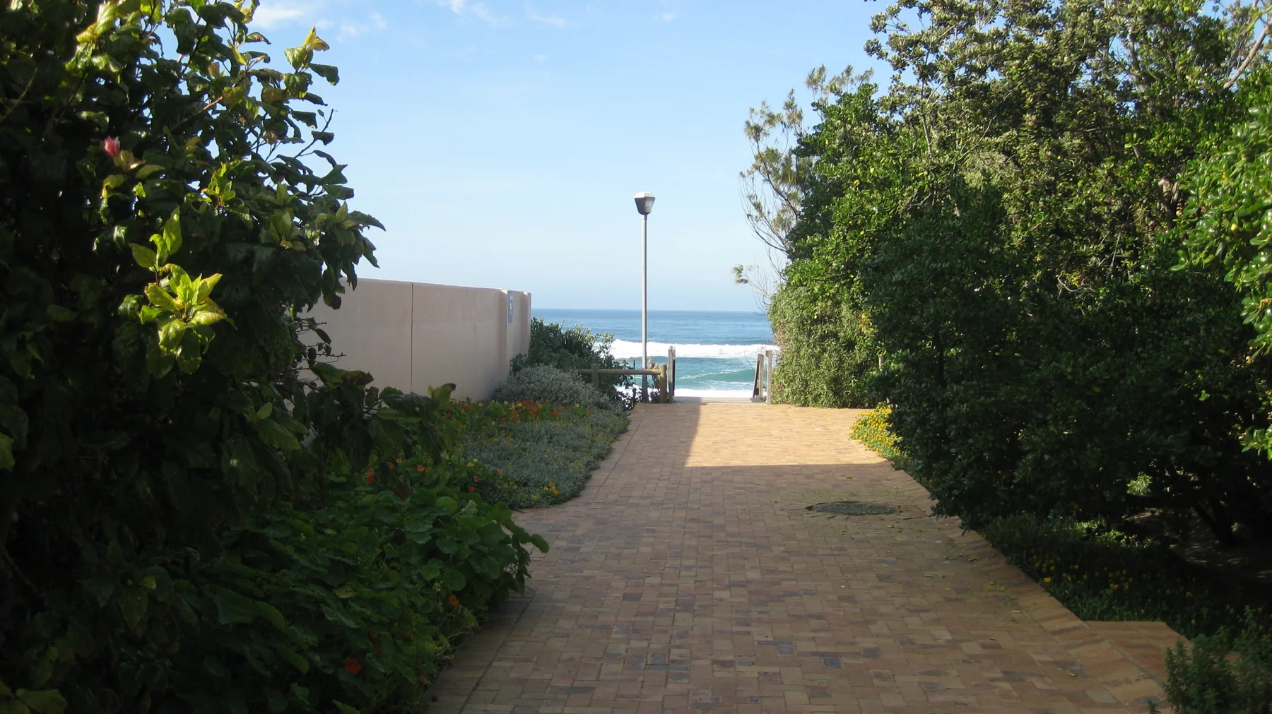 A paved walkway passes green hedges toward visible sea water.