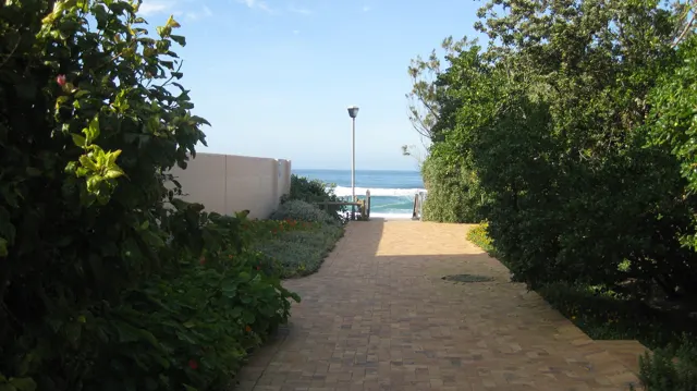A paved walkway passes green hedges toward visible sea water.