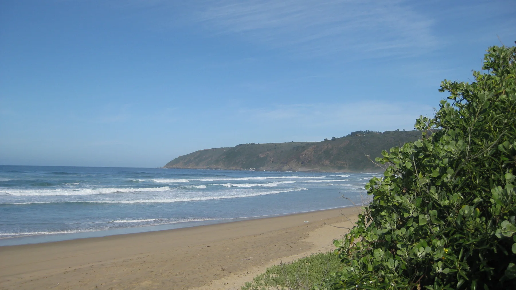 Waves wash a broad beach below a green headland.