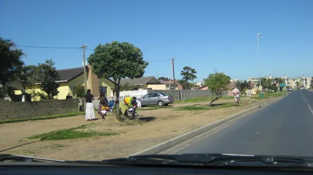 People stand on a sandy verge near houses and parked vehicles.