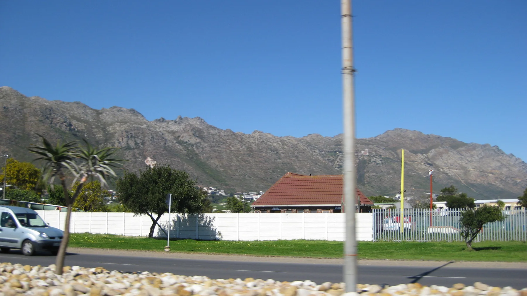 A red-roofed building stands behind a white wall below mountains.