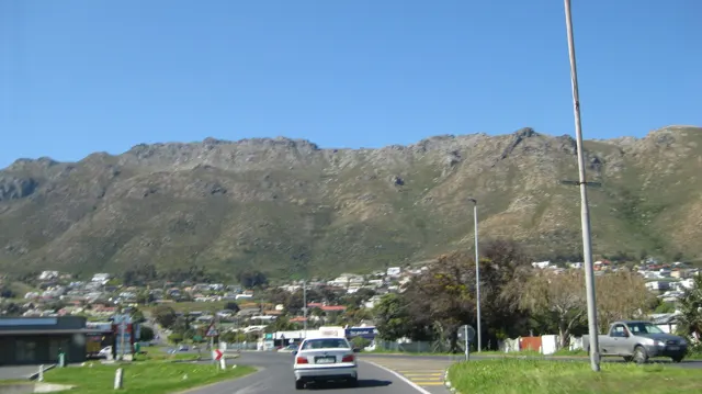 Cars follow a town road beneath a rugged mountain wall.
