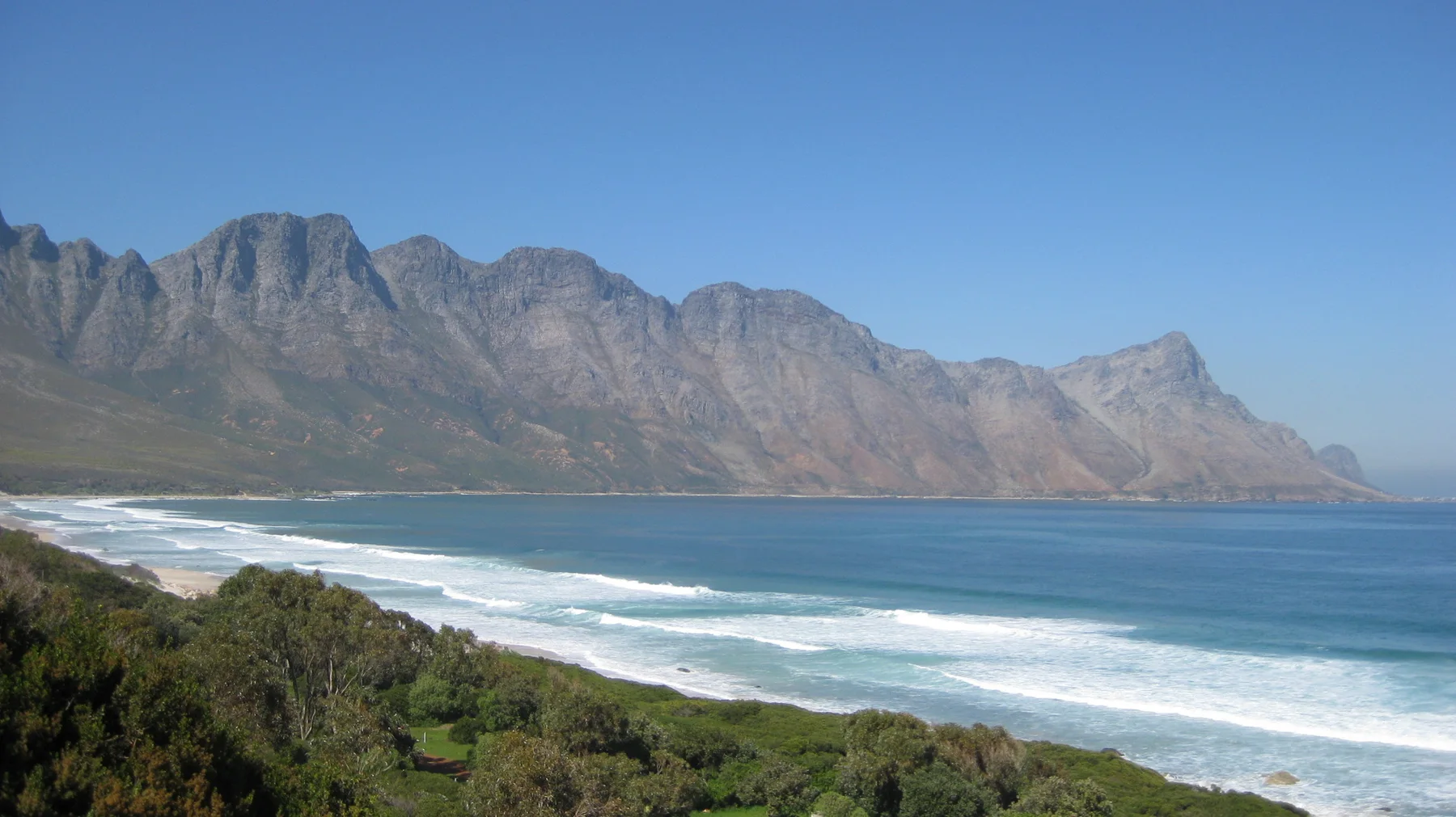 A long mountain wall rises beyond blue water and breaking waves.