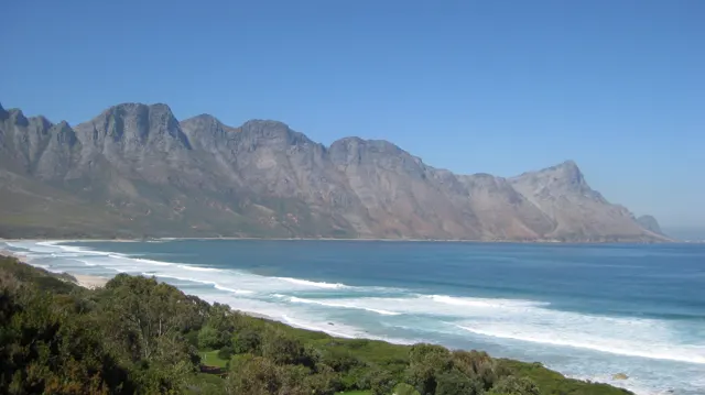 A long mountain wall rises beyond blue water and breaking waves.