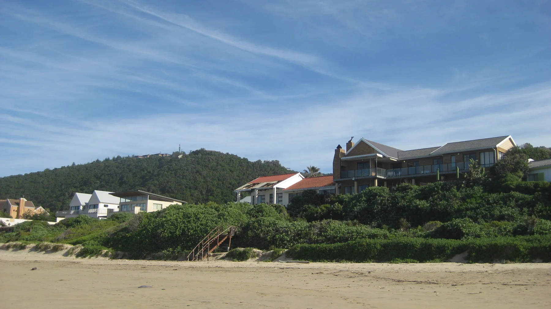 Houses stand above pale sand behind dense coastal plants.