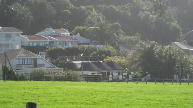 Houses sit below a wooded slope beyond a bright green field.