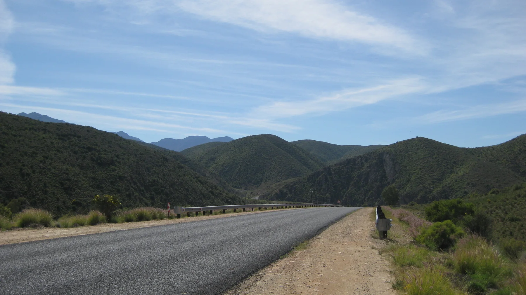 A person stands on the gravel verge of a road between green hills.
