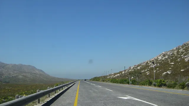 A straight highway runs between rocky green slopes beneath blue sky.