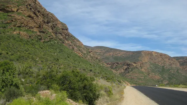 A paved road bends beside a reddish rock face and green shrubs.