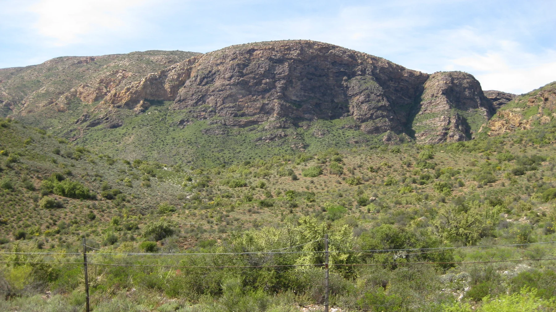 A broken brown cliff rises above a green valley slope.