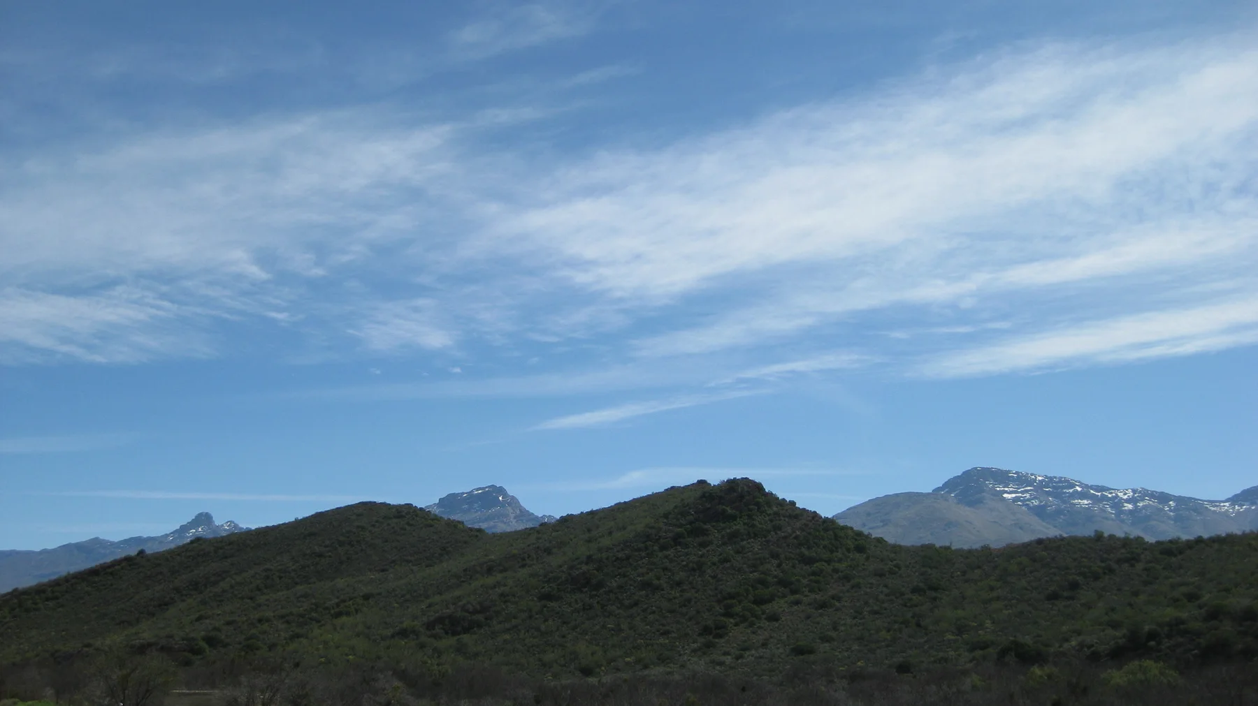 Thin white clouds stretch above rolling green hills and distant peaks.