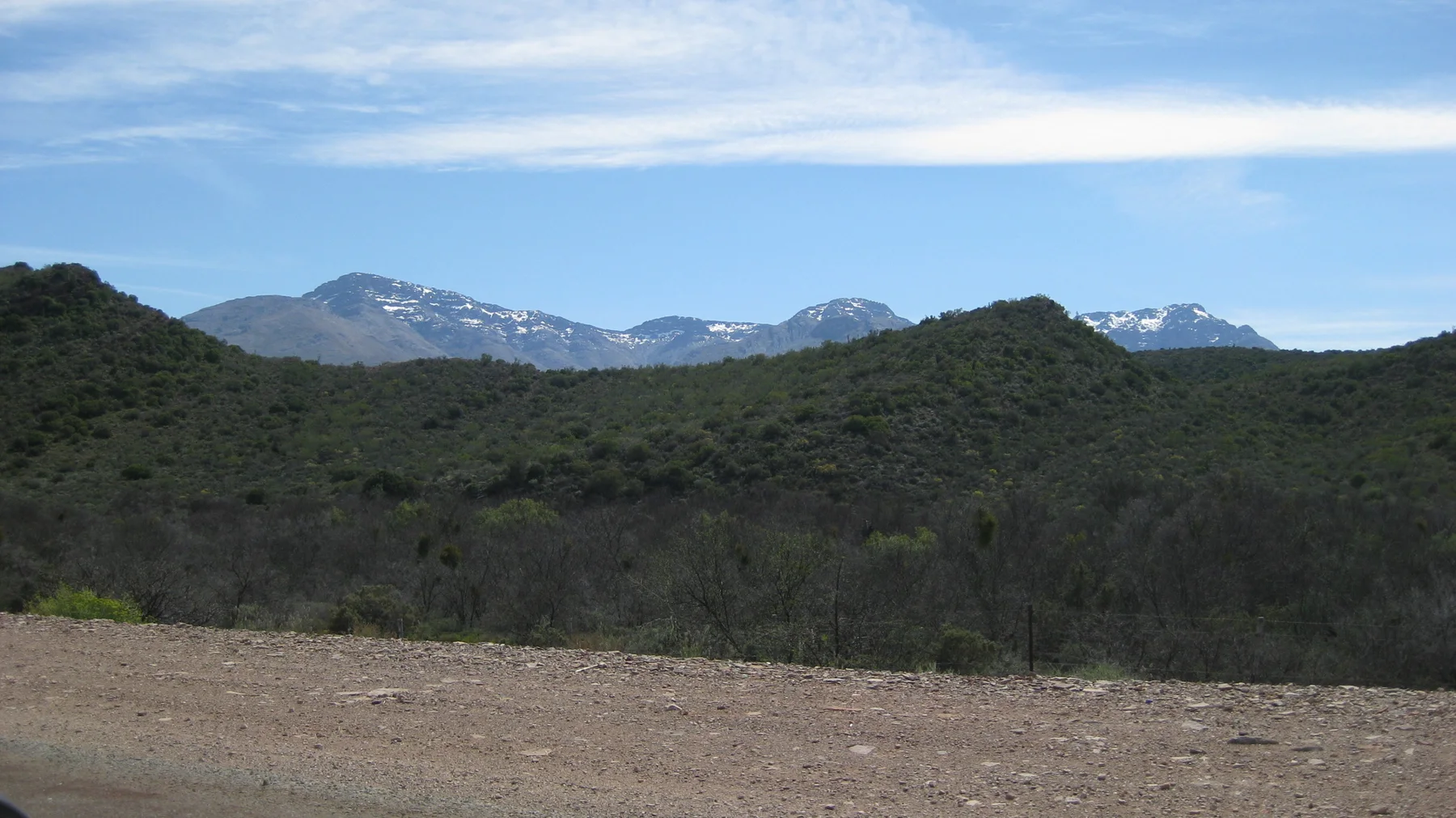 A gravel verge borders layered green hills and distant pale peaks.