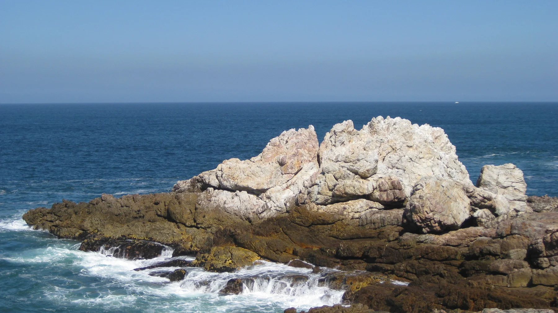 A large pale rock formation rises above dark shoreline rocks and surf.