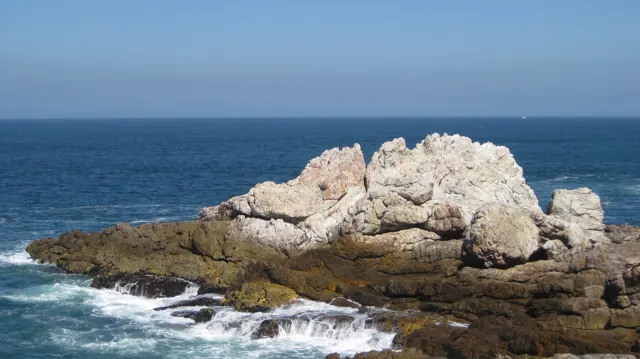 A large pale rock formation rises above dark shoreline rocks and surf.