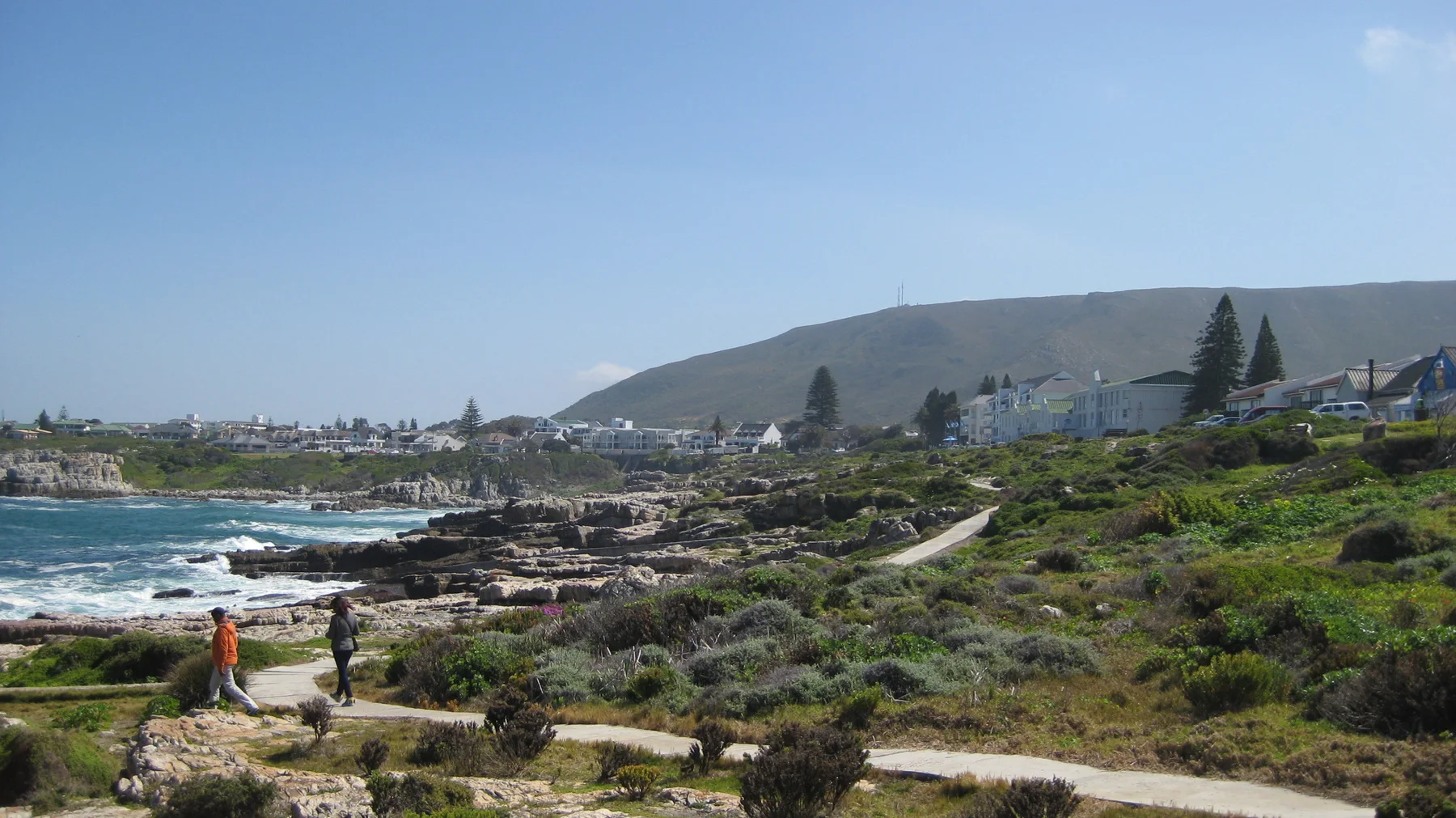 People follow a path above a rocky coastline and scattered buildings.