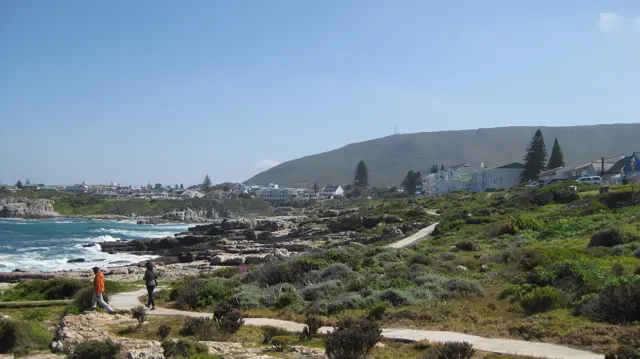 People follow a path above a rocky coastline and scattered buildings.