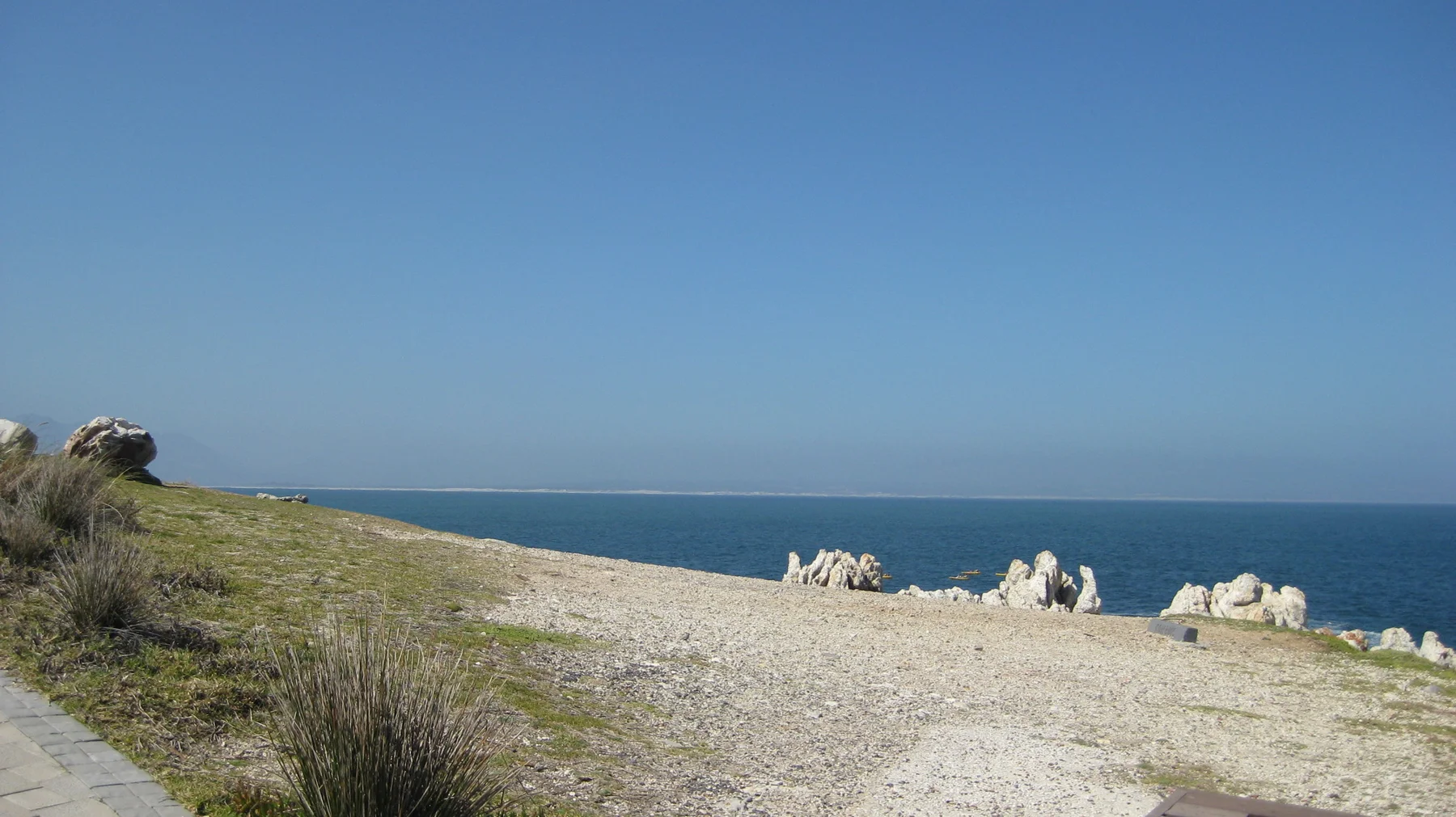 A sandy lookout faces blue sea beyond low rocks and grass.