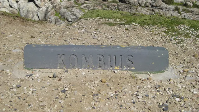 A plaque marked Kombuis is set into sandy ground beside rocks.