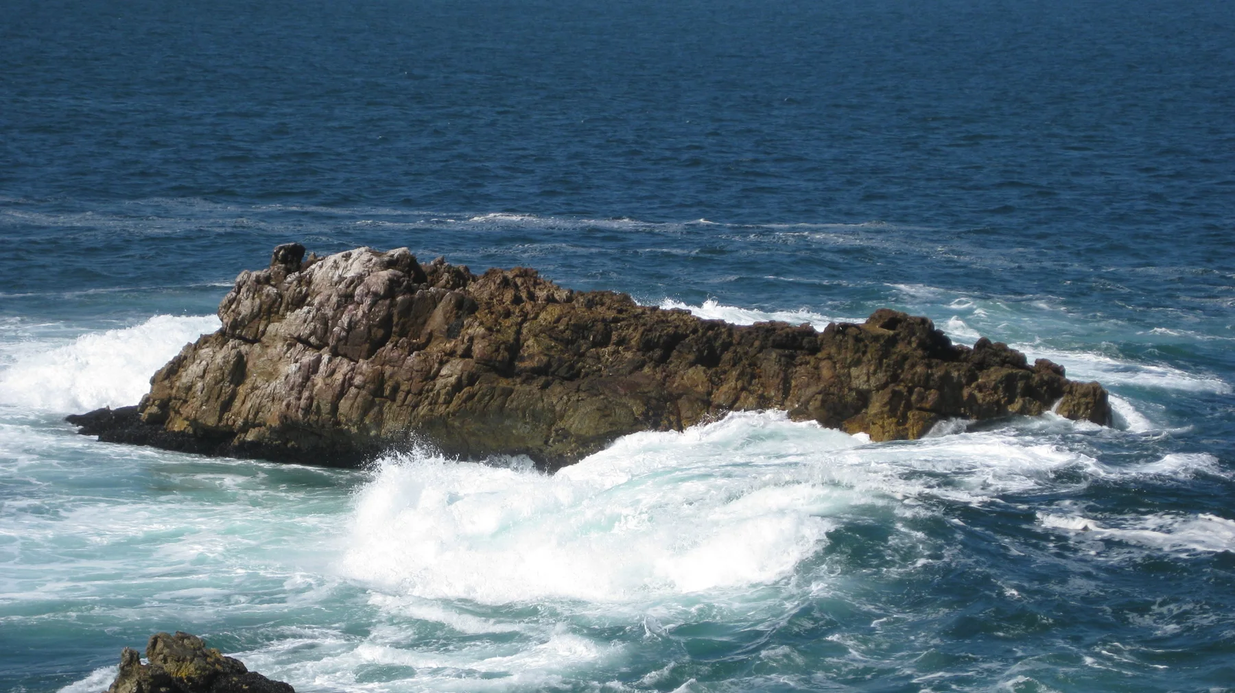 A white wave breaks against a low offshore outcrop.