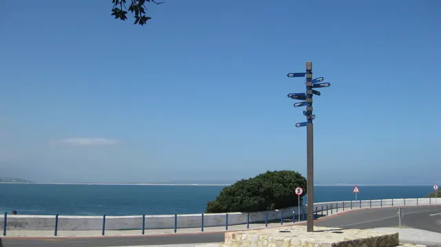 A tall signpost stands above a railing-lined seaside promenade.