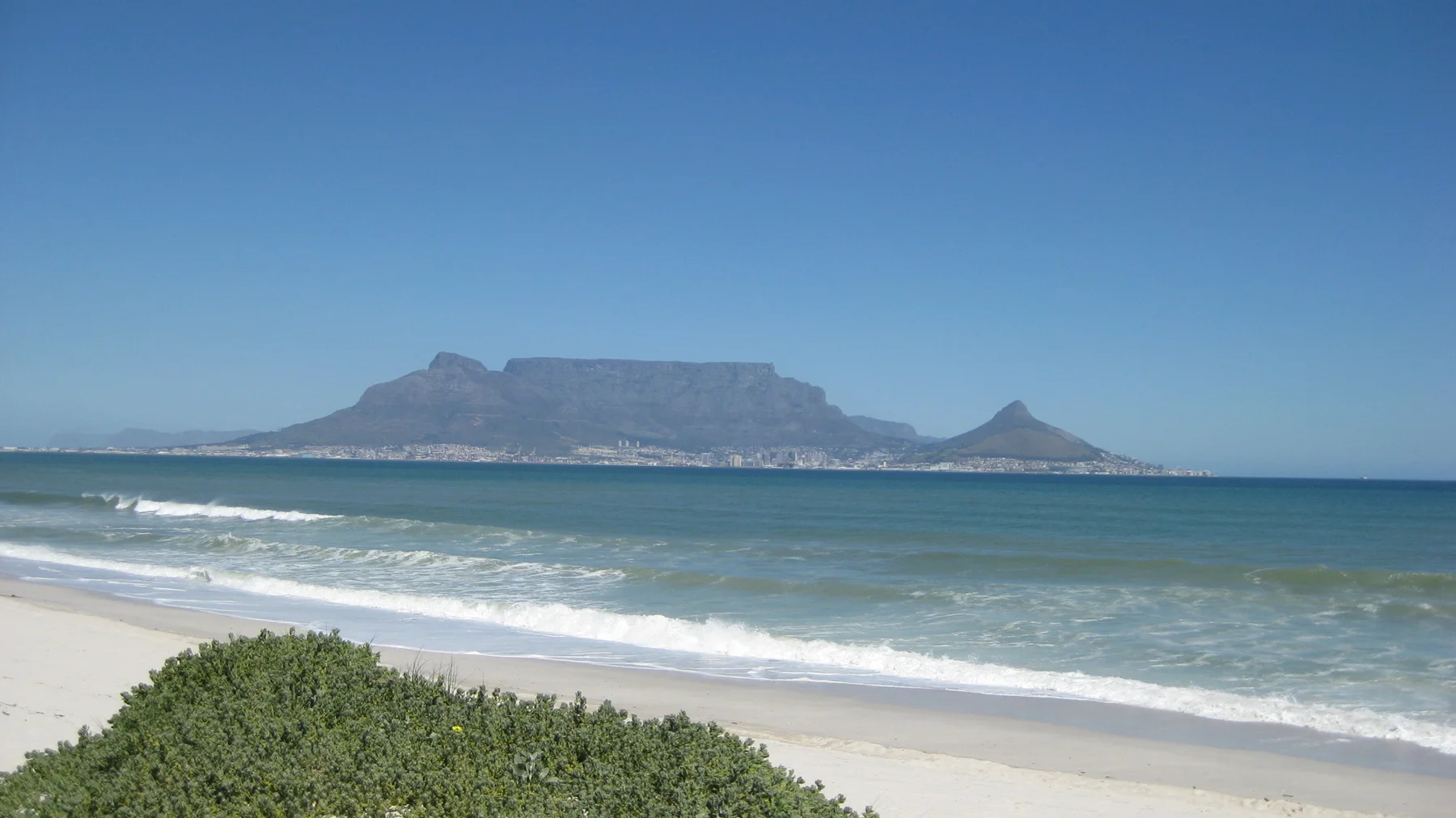 A flat-topped mountain rises across blue water beyond white surf.