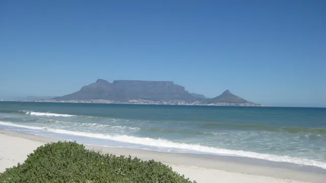 A flat-topped mountain rises across blue water beyond white surf.
